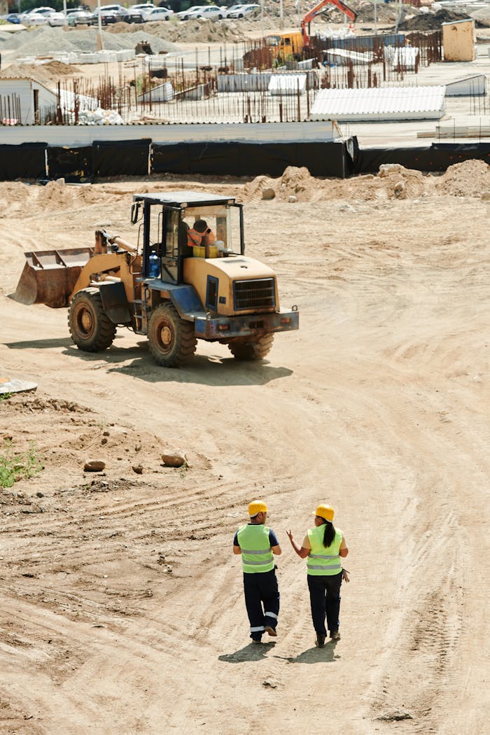 services-06 Construction workers in safety gear walking at a busy site with a backhoe in operation.