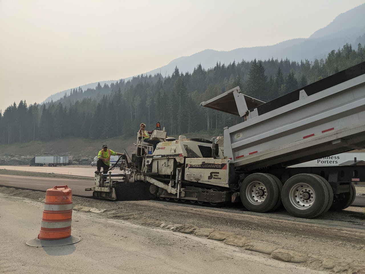 about-bg Road workers paving a highway amidst mountainous forest scenery.